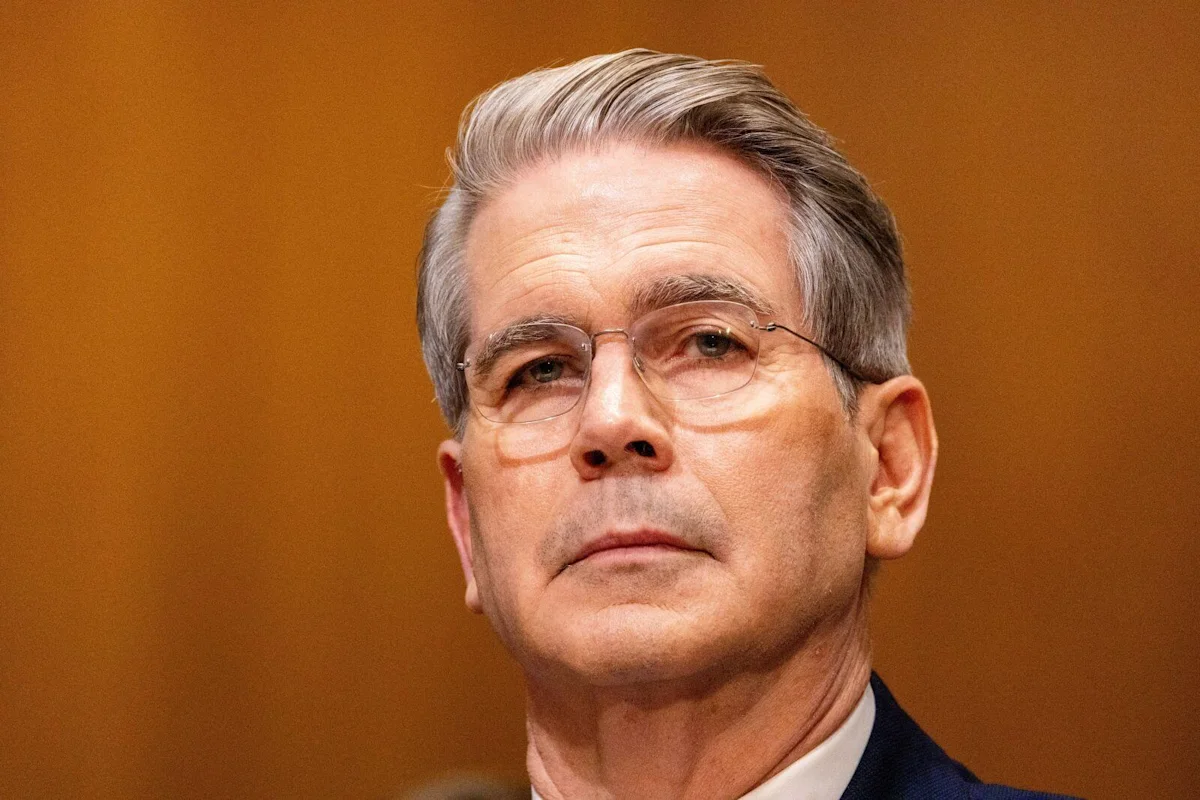 Gray-haired man wearing glasses and a dark suit against a warm brown background