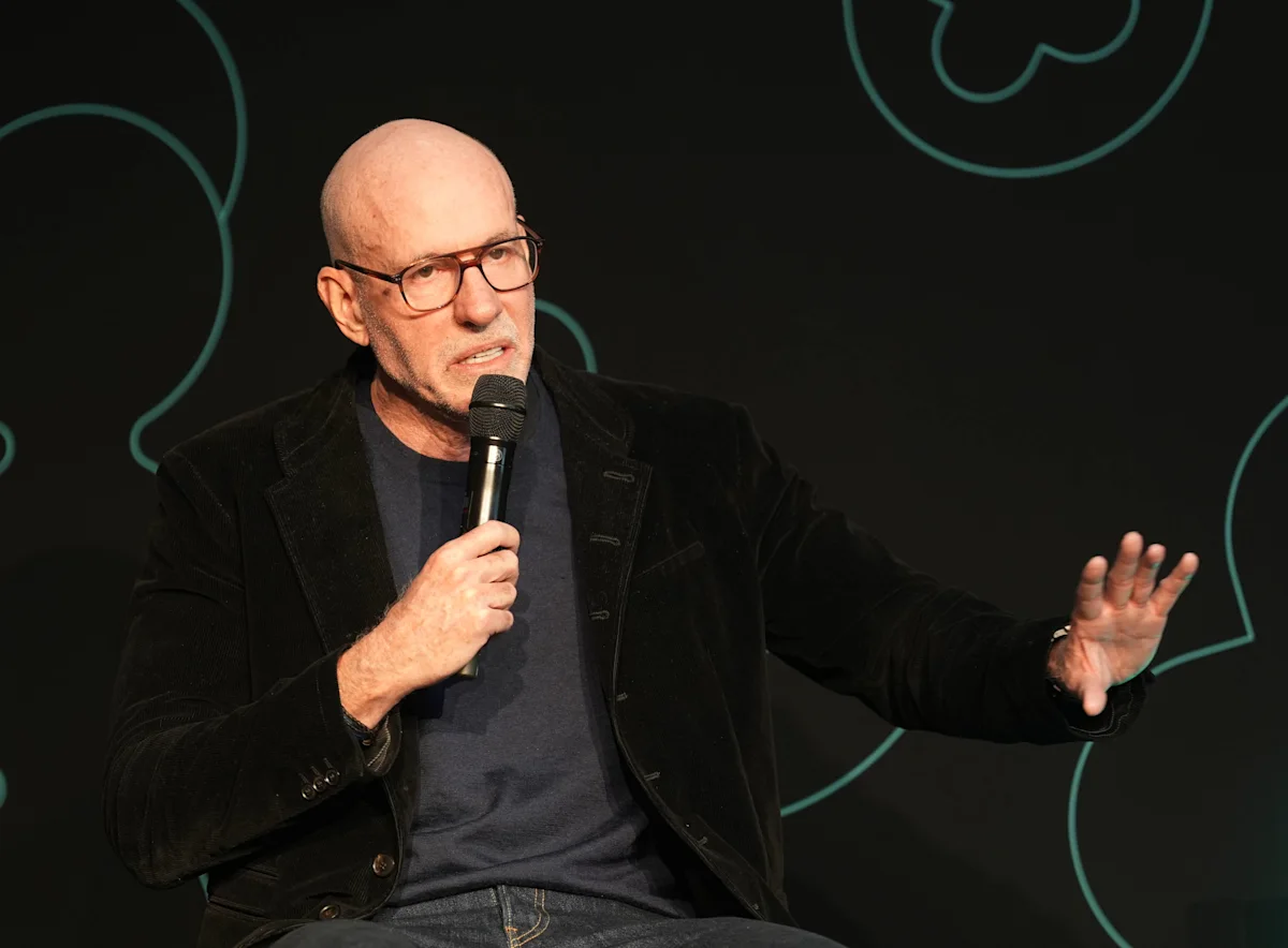 Bald man wearing glasses speaks into a microphone while gesturing onstage.