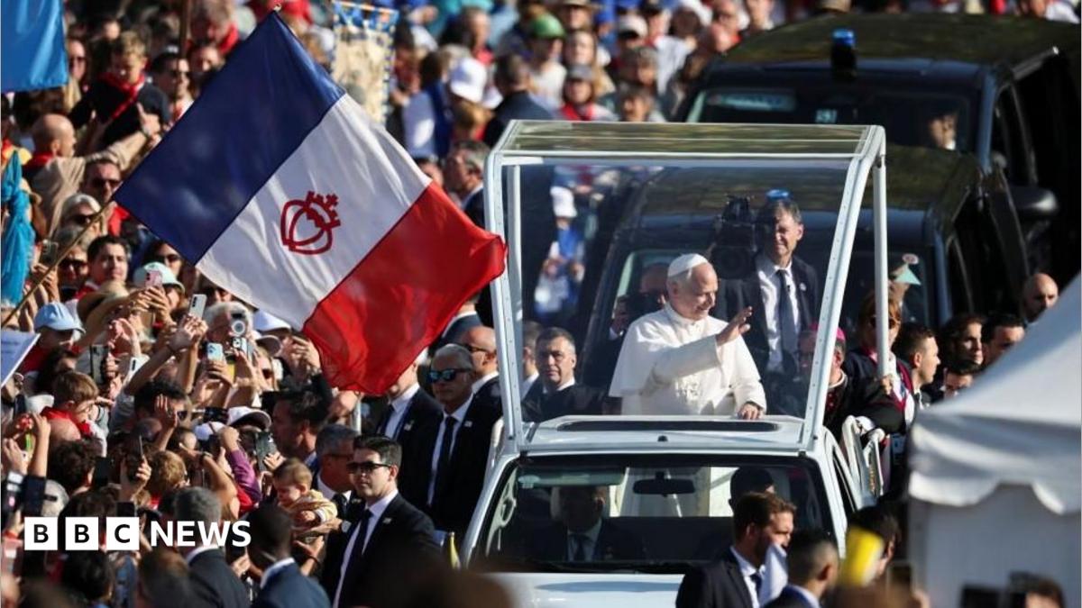 A white-robed man waves from a clear-sided vehicle amid a large crowd and a French flag.
