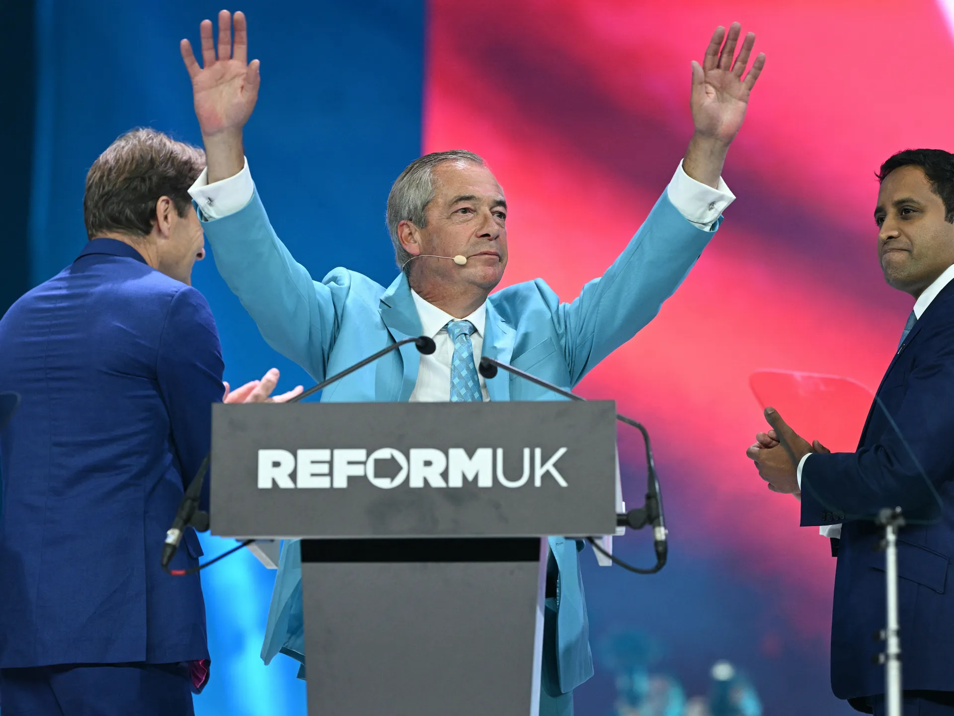 Speaker in light blue suit raises both hands at a Reform UK podium, flanked by two men.