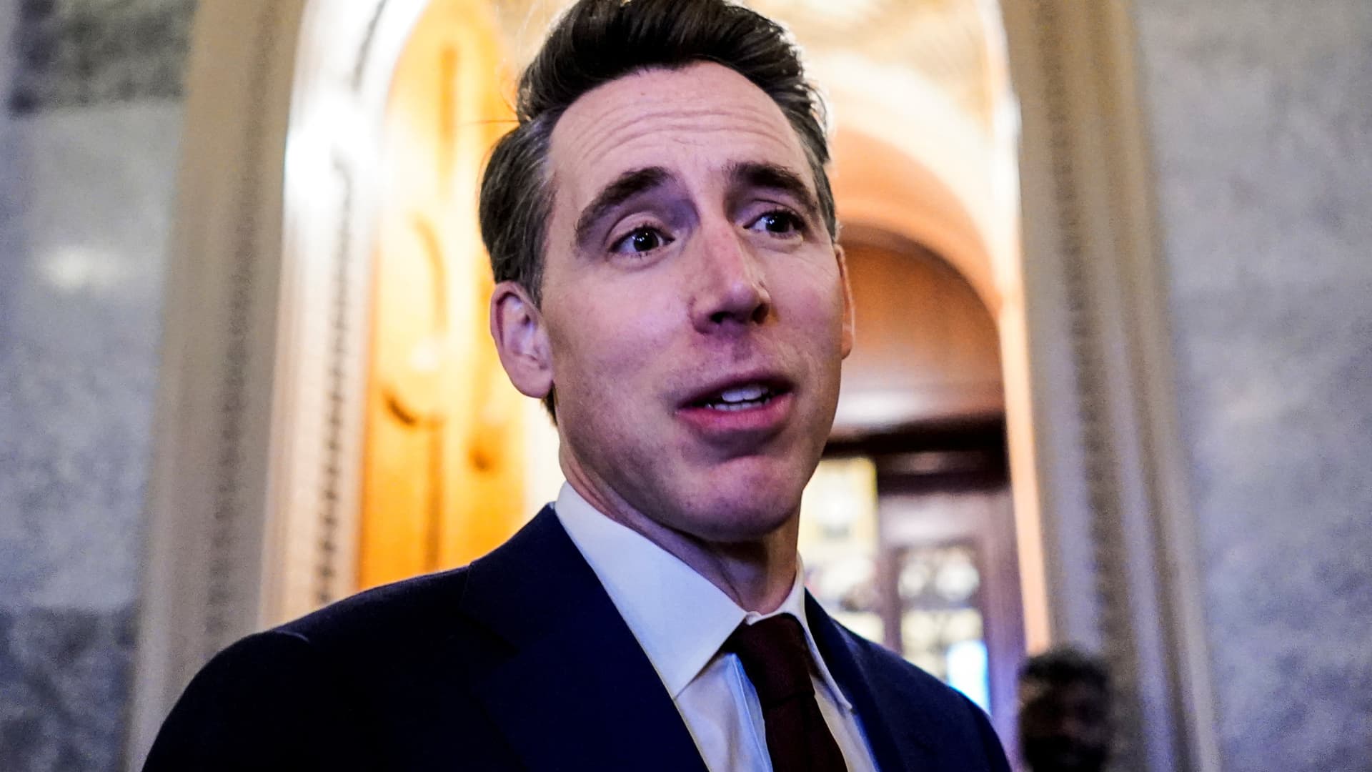 Man in a dark suit and tie stands before a brightly lit arched interior.