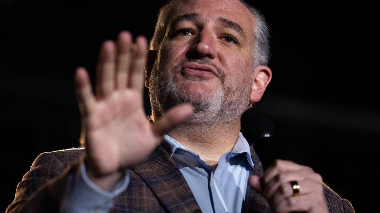A man in a checked blazer speaks into a microphone and raises an open hand against a dark background.