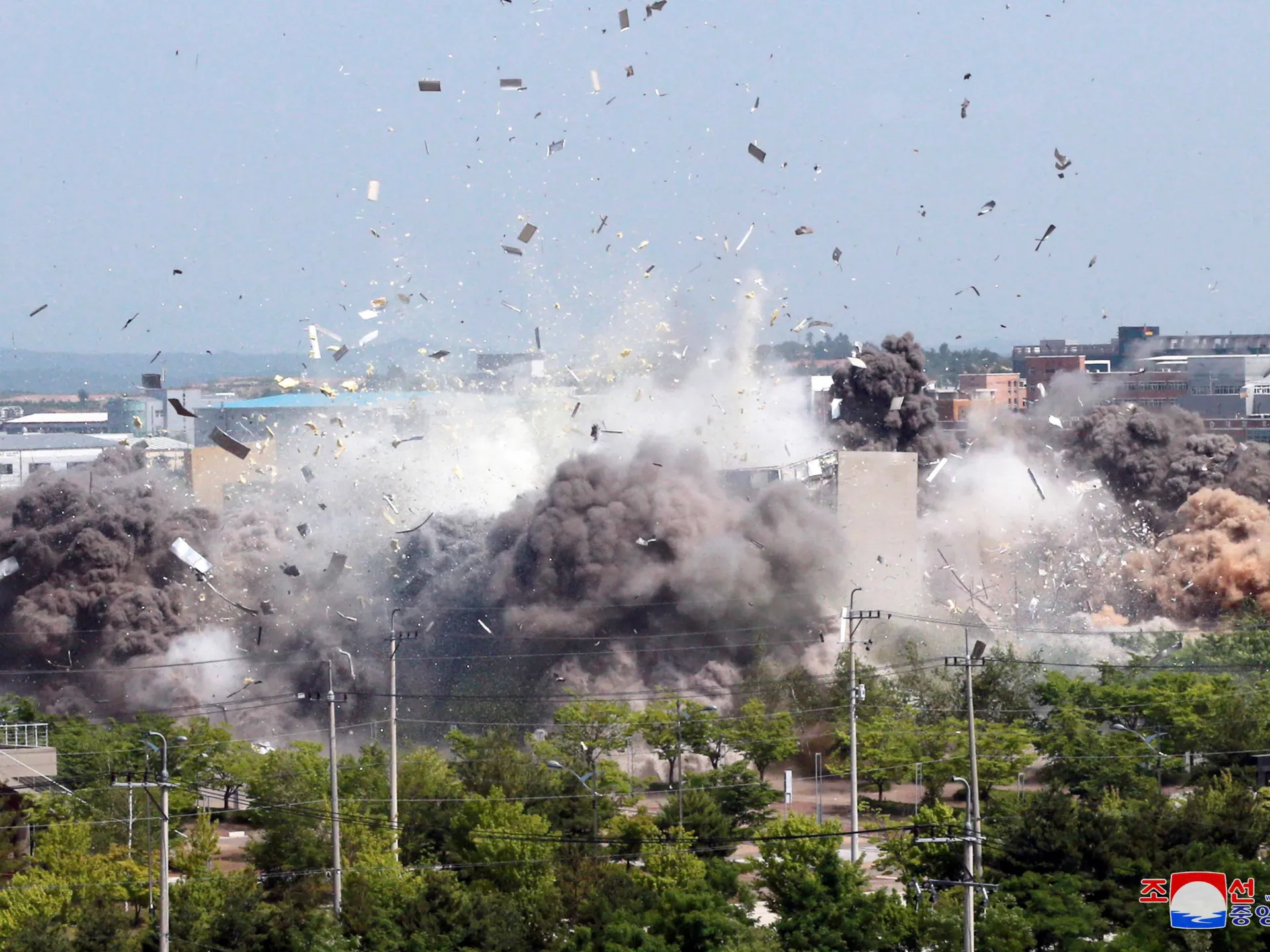 Large explosion sends thick smoke, debris, and dust above a cityscape and tree-lined street.