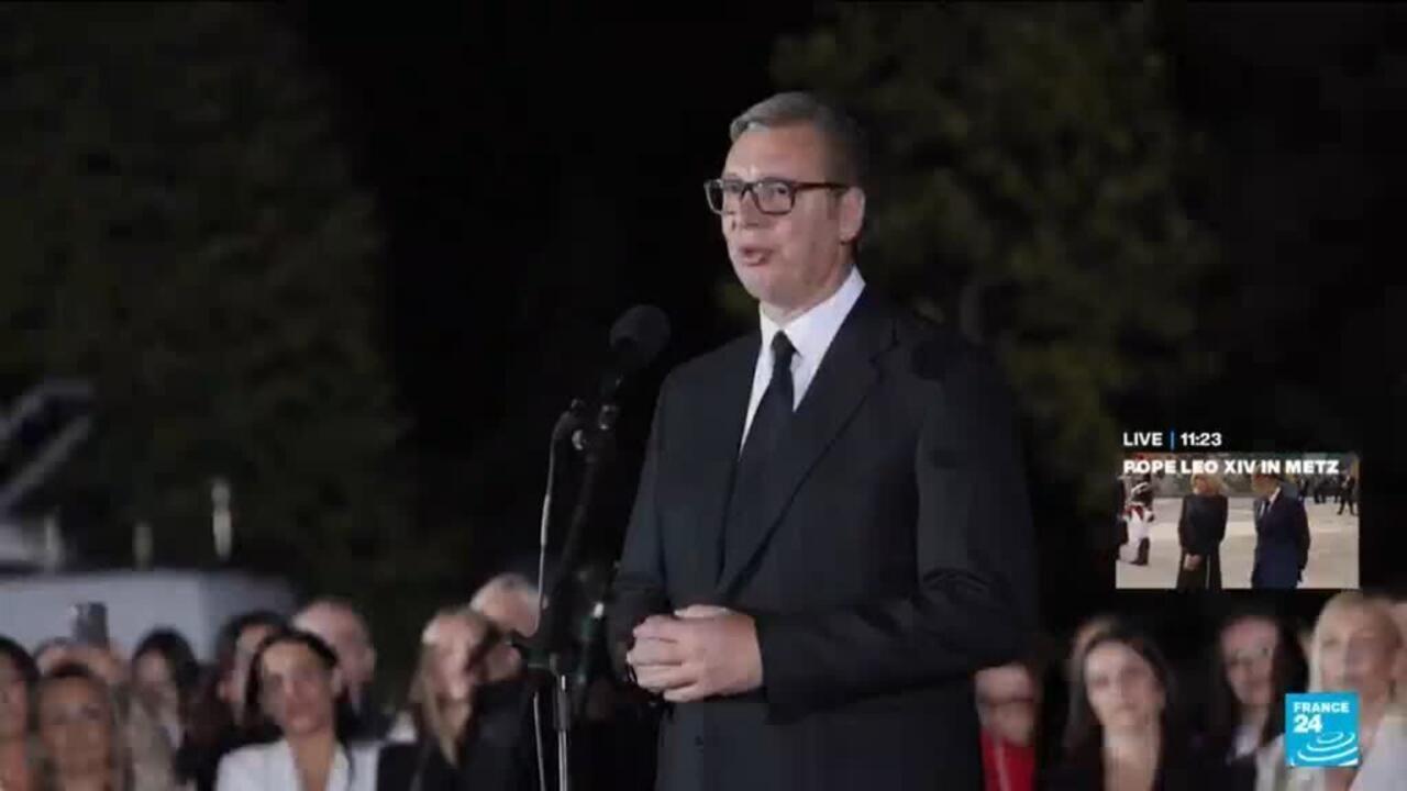 A suited man with glasses speaks at a microphone before a seated audience outdoors at night.