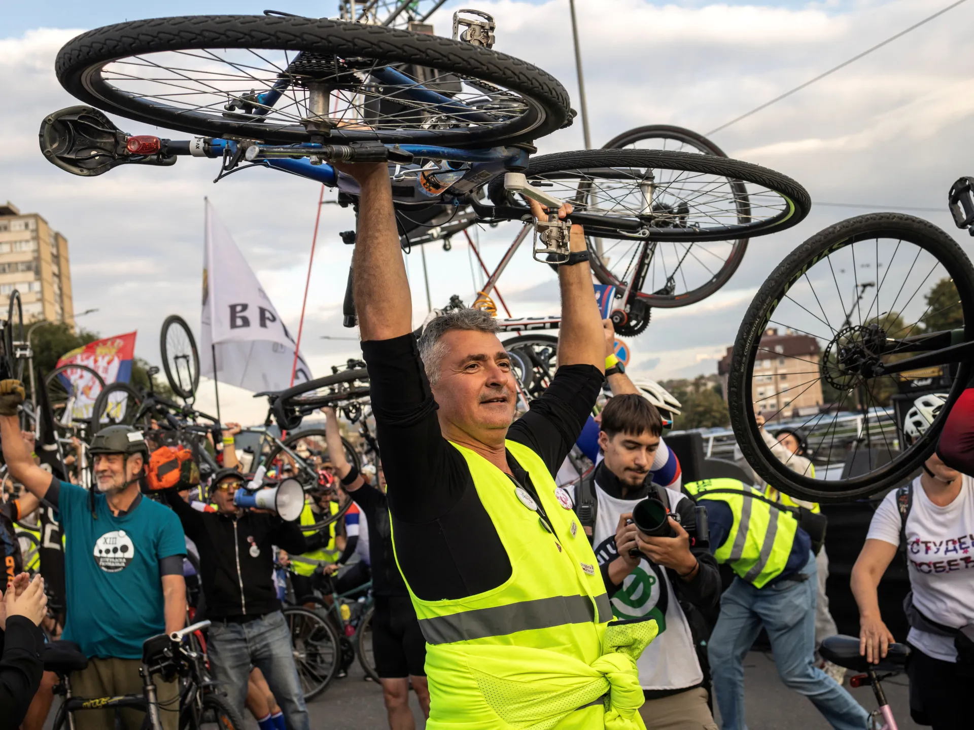 A man in a fluorescent yellow vest raises a bicycle above a crowd of cyclists, some wearing helmets and similar vests.