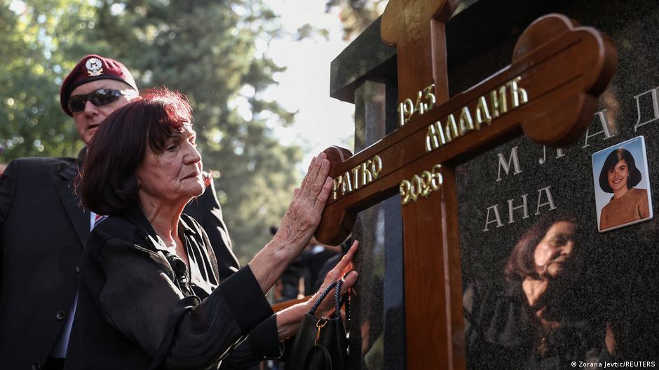 Woman touches a memorial cross beside a photograph while a uniformed man stands behind her.