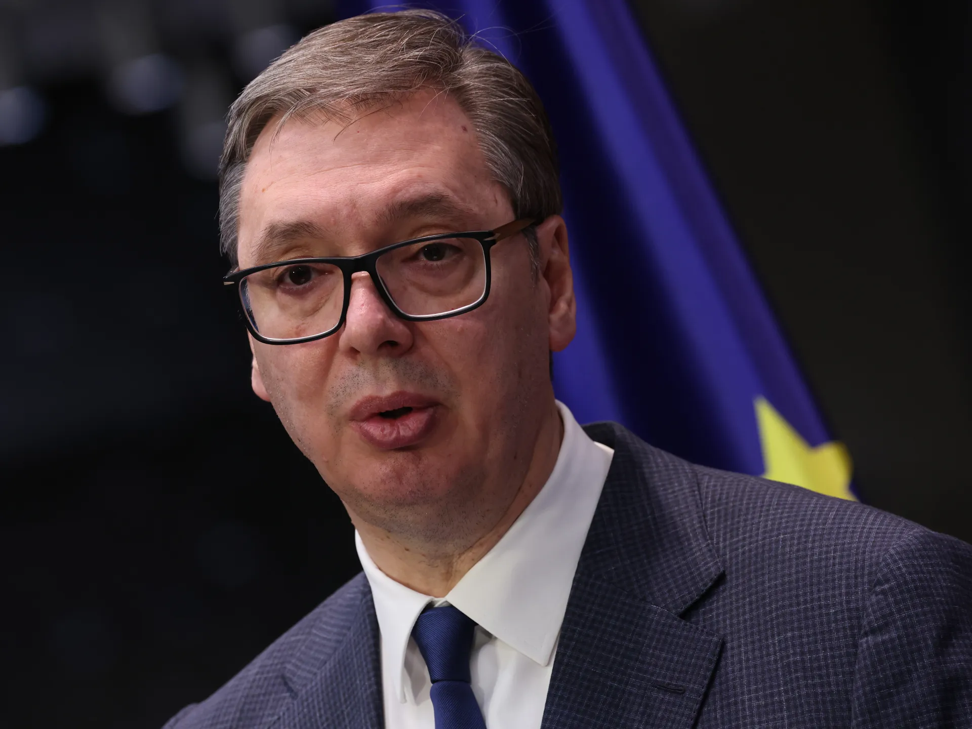 Close-up of a bespectacled man in a suit speaking against a dark background with a blue flag.