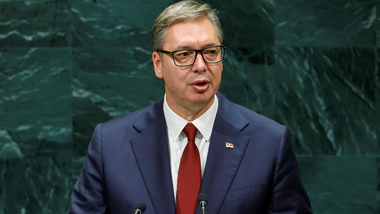 A man in glasses and a navy suit speaks at a podium against a dark green marble wall.