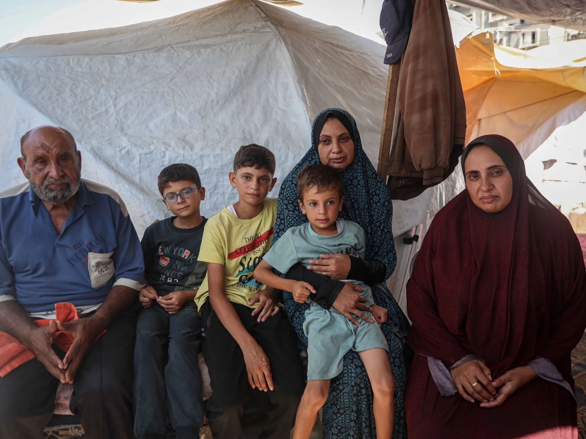 Six people, including three children, sit together inside a makeshift tent with two women and a man.