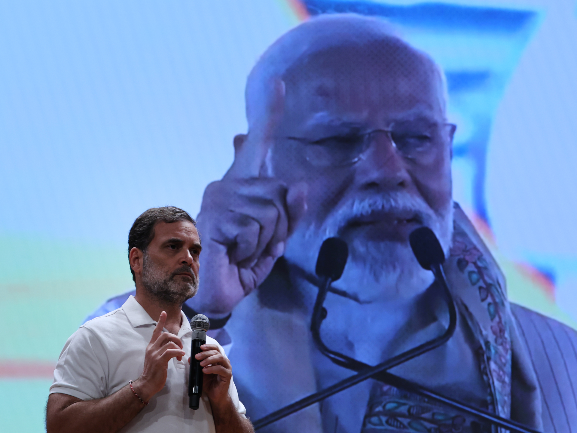 A man speaks into a microphone before a large projected image of an older man at a microphone.