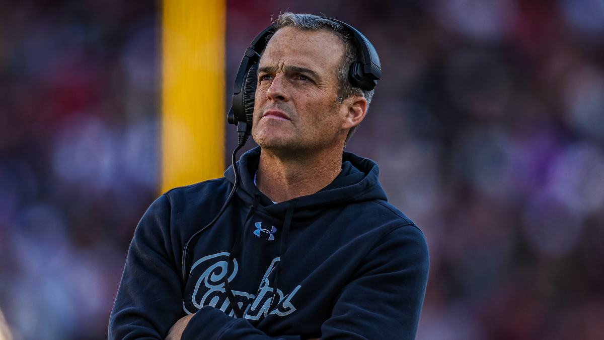 A man in a dark South Carolina hoodie and headset stands with arms crossed at a football game.