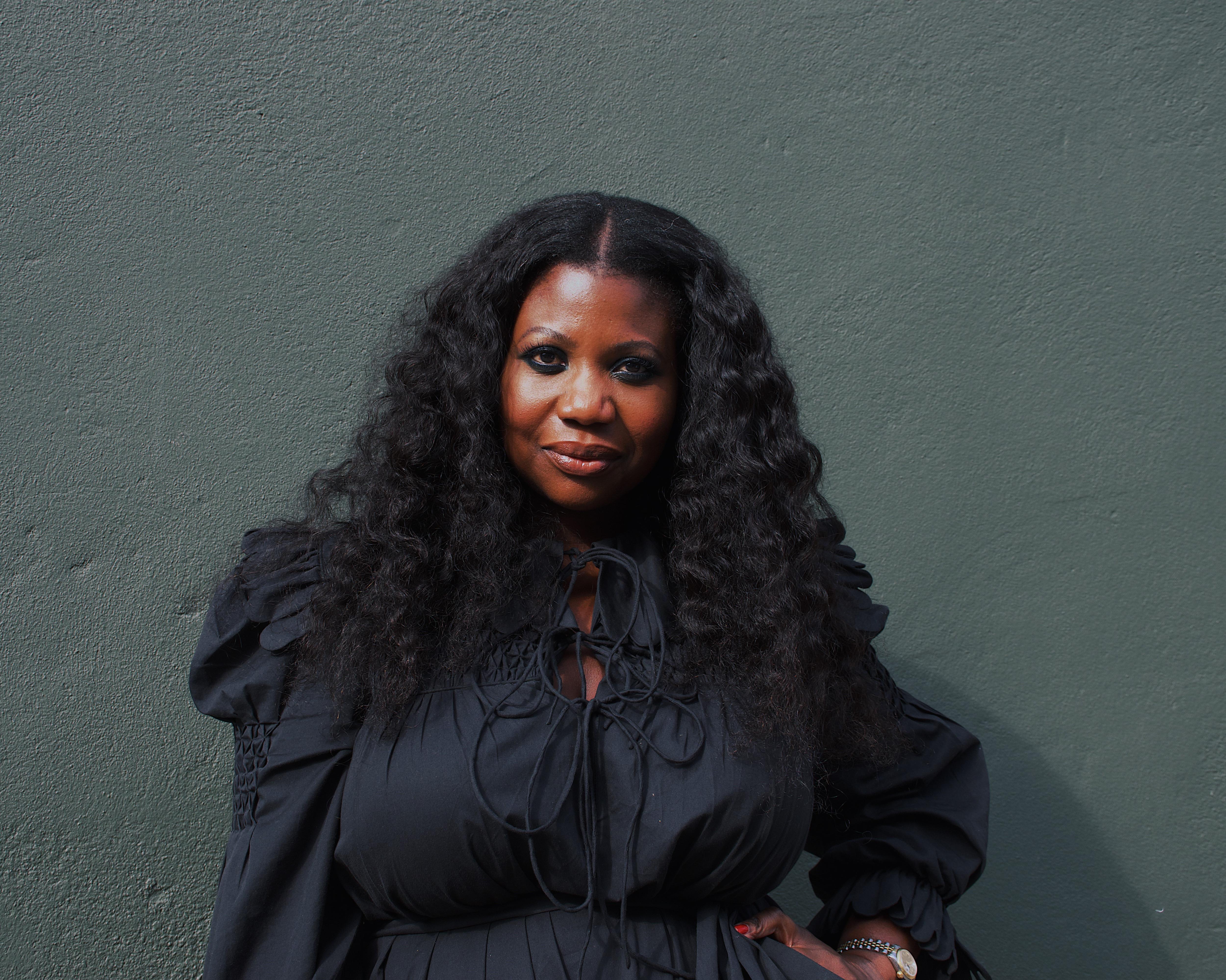 Portrait of a Black woman with long wavy hair wearing a black blouse against a textured green wall.