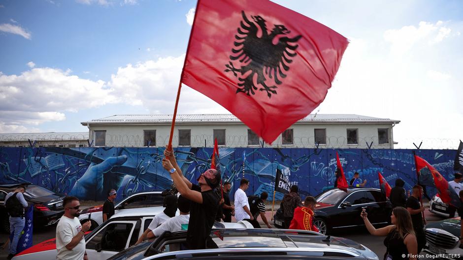 Crowd waving Albanian flags beside cars and a colorful mural beneath a bright sky