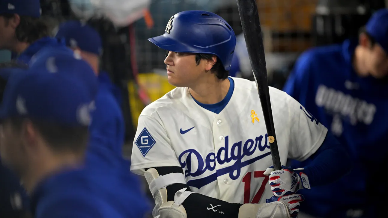Baseball player in Dodgers uniform and helmet holding a bat near the dugout