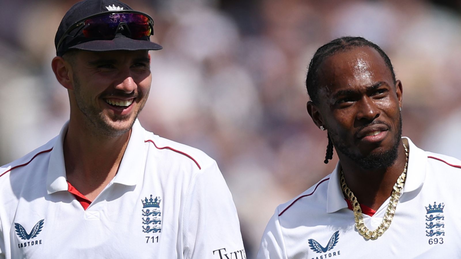 Two England cricketers in white shirts stand together, one smiling beneath a cap and sunglasses.