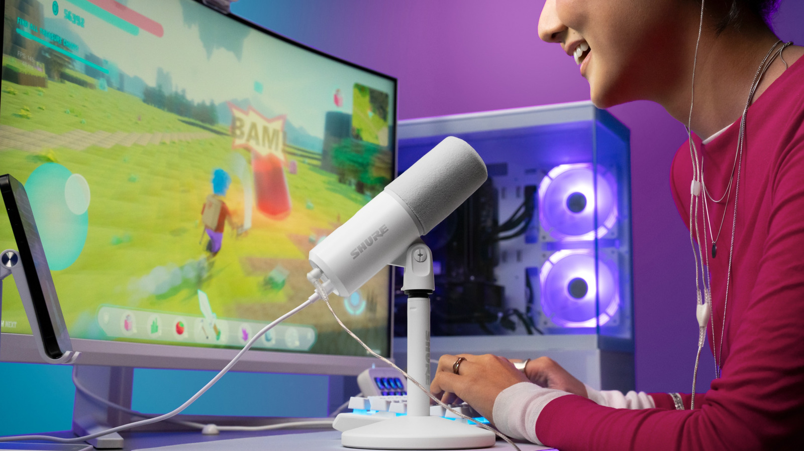 A person wearing a magenta top uses a white Shure microphone beside a gaming monitor and illuminated computer.