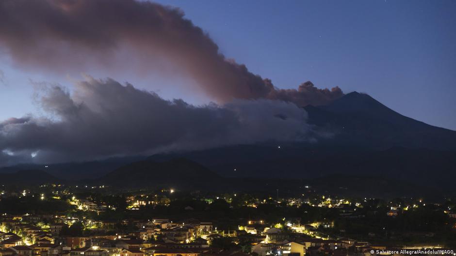 A large plume of ash and smoke billows from a mountain above a town at dusk.