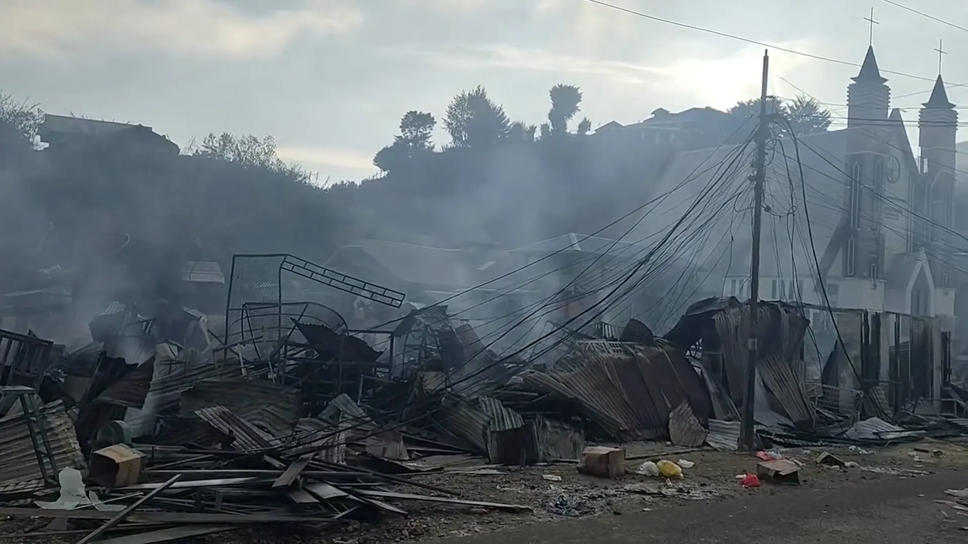 Charred market stalls smolder beneath tangled utility wires beside a church-like building.