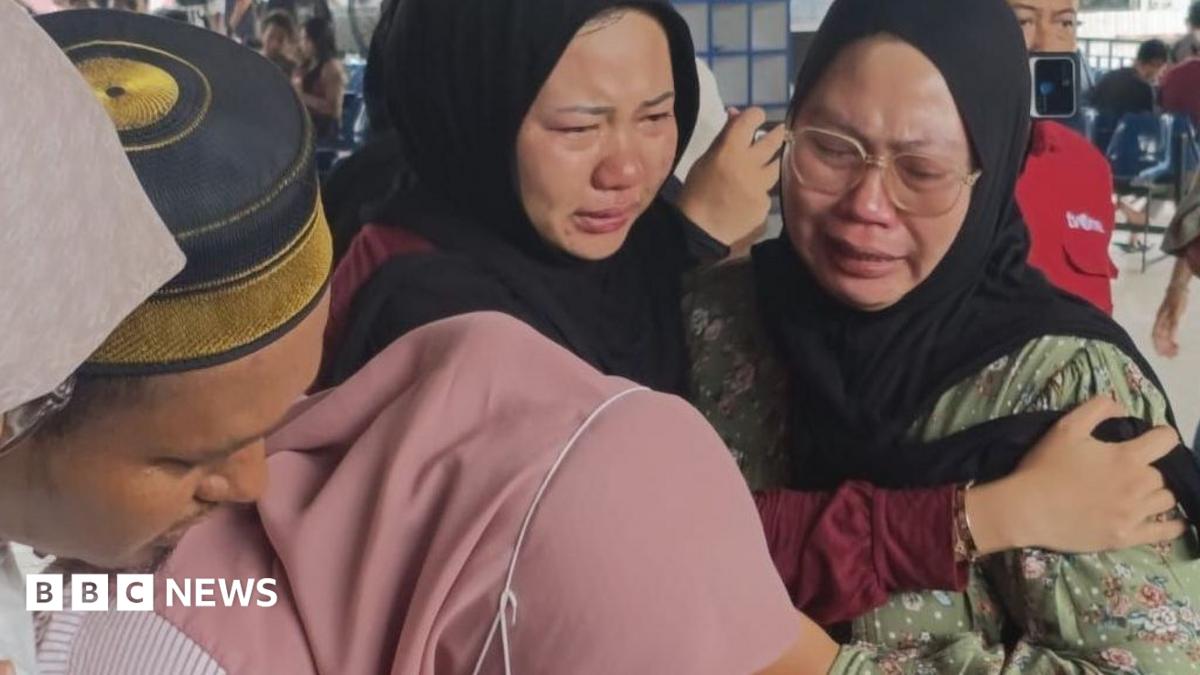 Two crying women in black headscarves are comforted amid a crowded indoor setting.