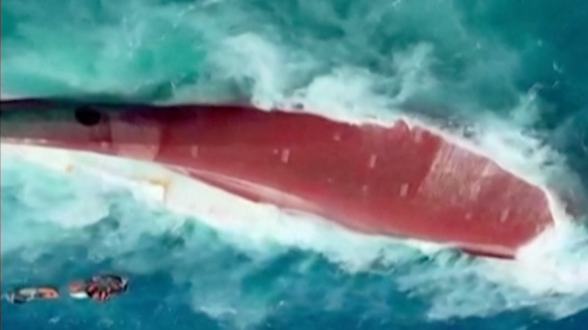 A large red vessel lies overturned in turbulent turquoise water as small boats approach.