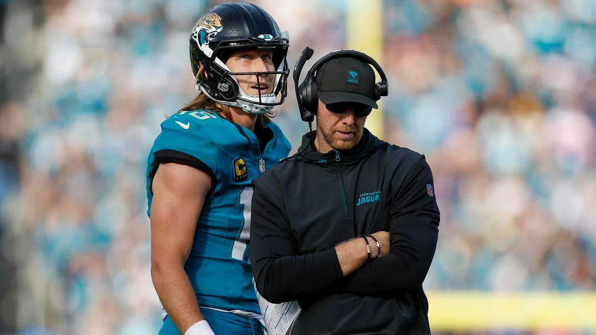 Football player in helmet stands beside a coach wearing a headset on the sideline.