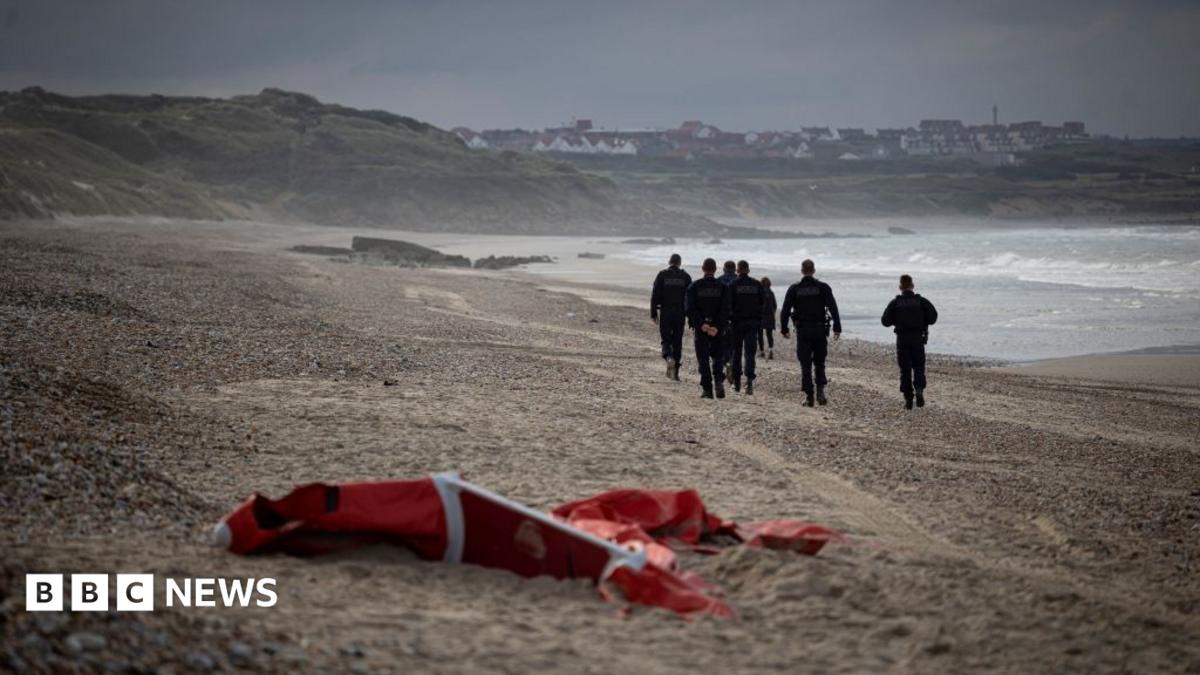 Several people in dark uniforms walk along a pebbled beach as waves break nearby; red equipment lies in the foreground.