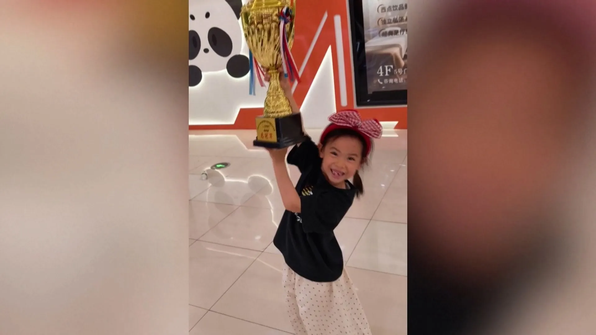 A smiling young girl in a black outfit raises a gold trophy indoors.