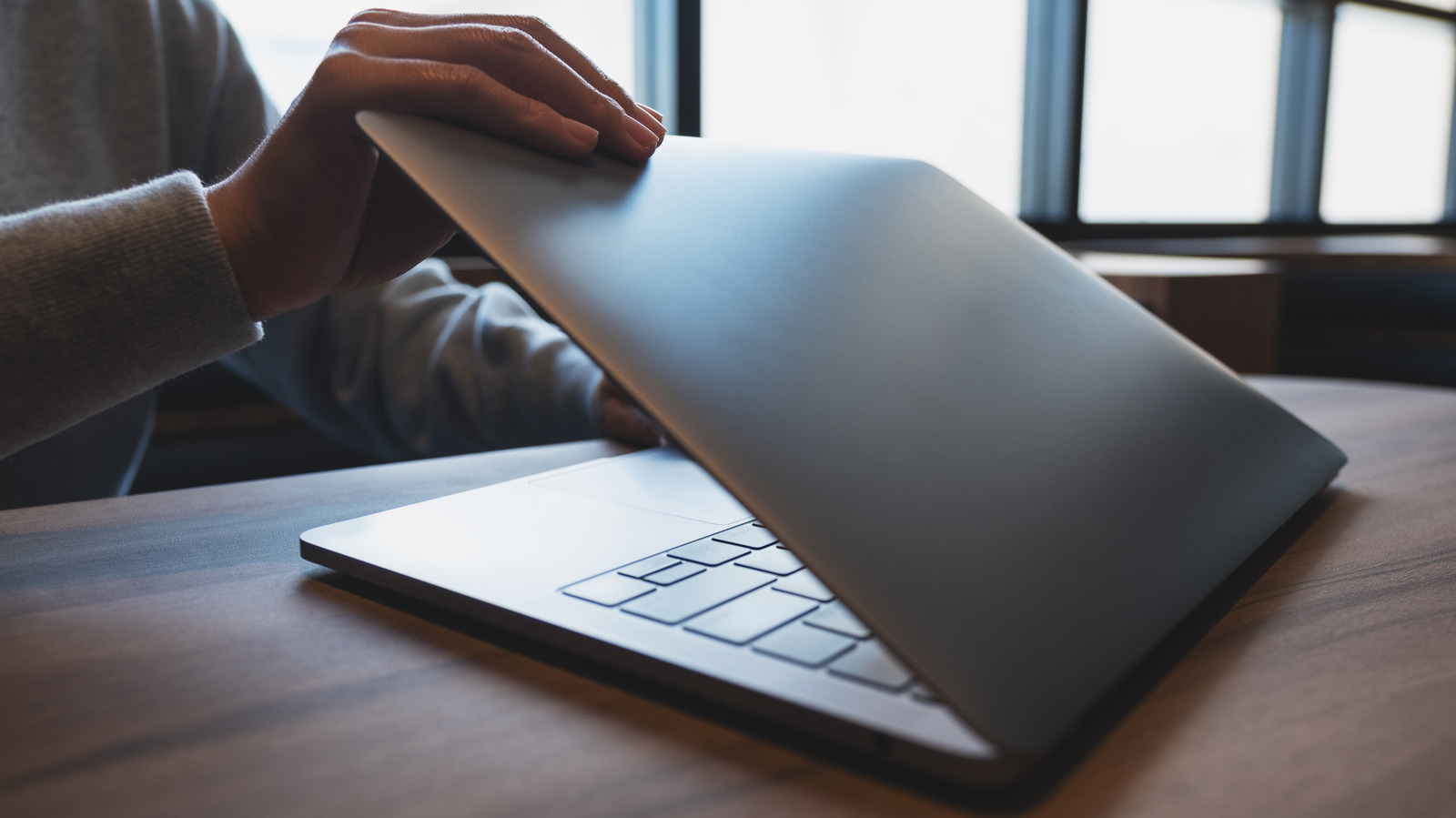 A person holds open a laptop on a wooden desk beside a bright window.