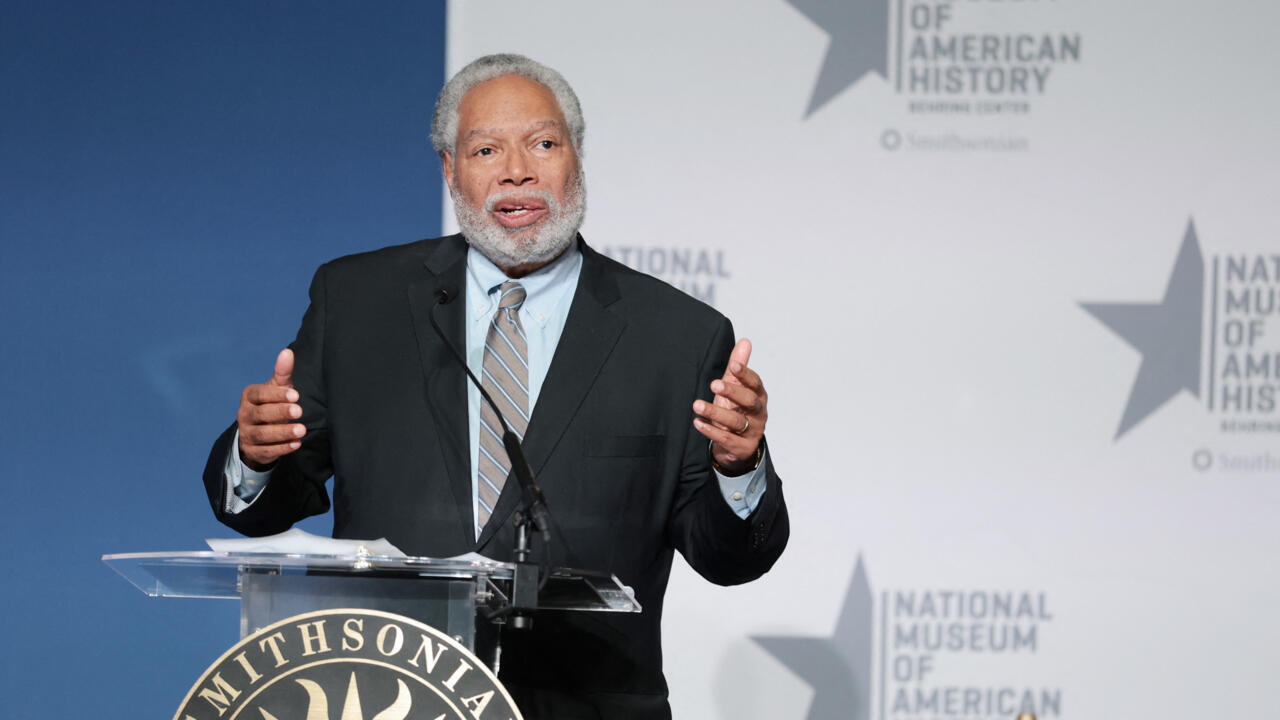 A man in a suit gestures while speaking at a Smithsonian podium.