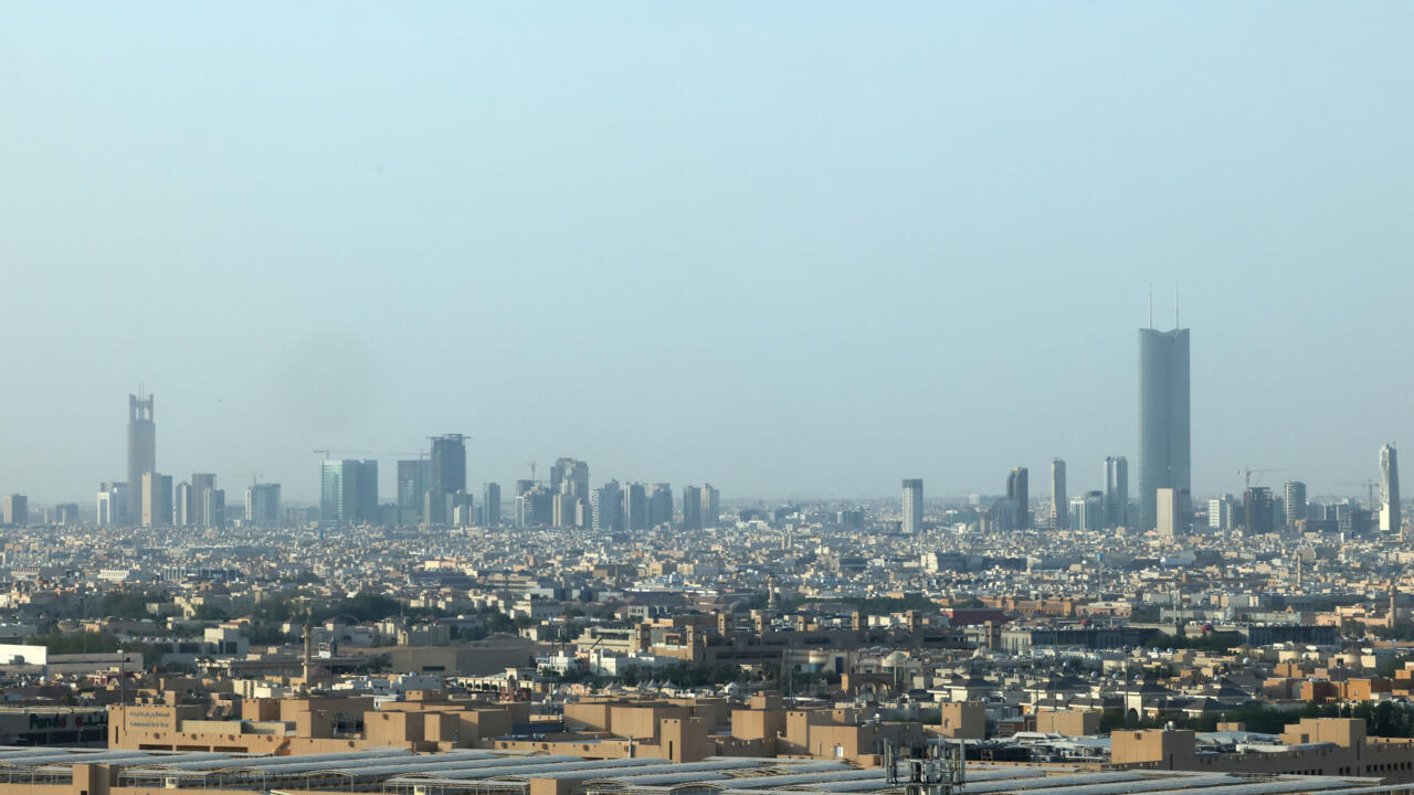 Wide city skyline with dense low-rise buildings and several tall towers beneath a hazy blue sky.