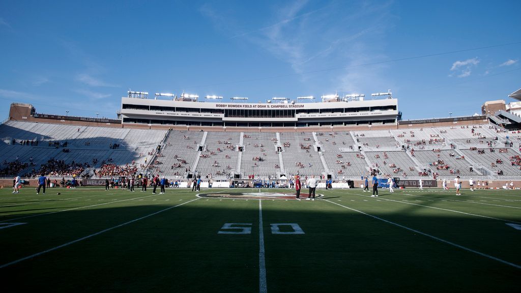 Football stadium viewed from the field, with empty bleachers, scattered spectators, and a clear blue sky.