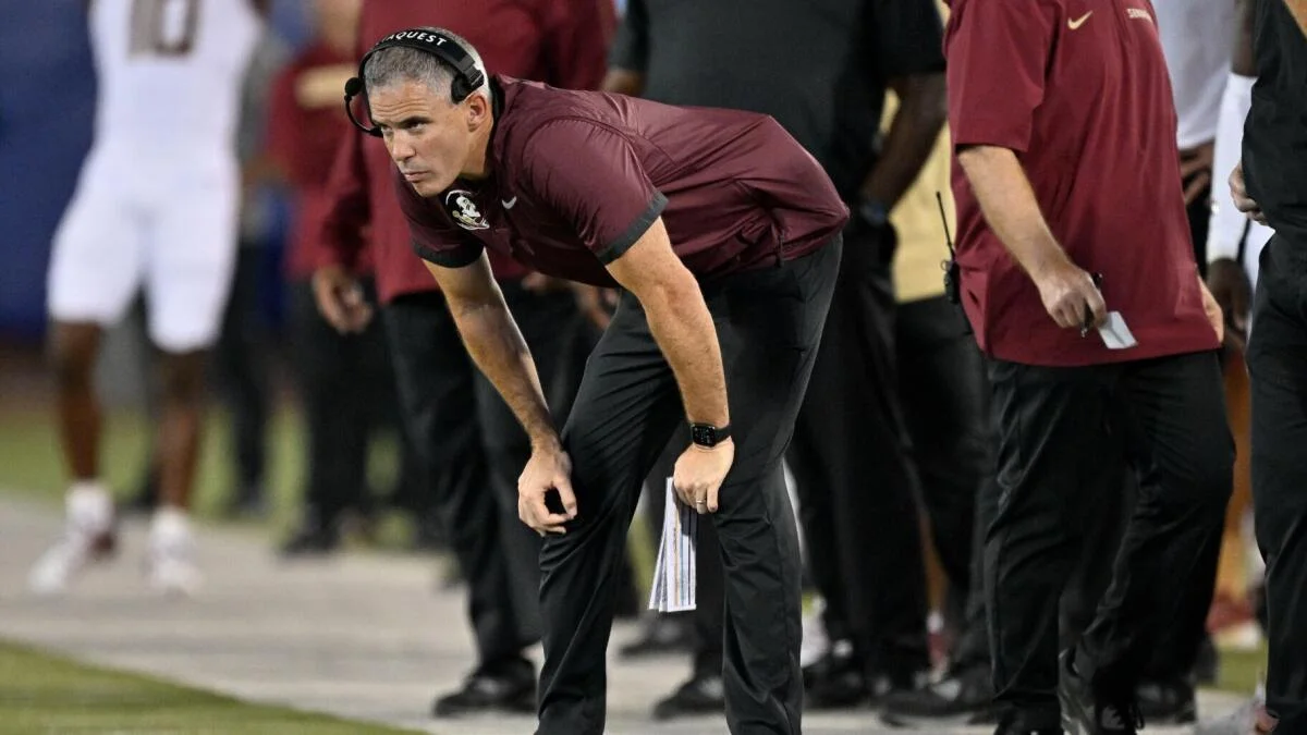 Maroon-clad football coach wearing a headset bends forward near the sideline during a game.