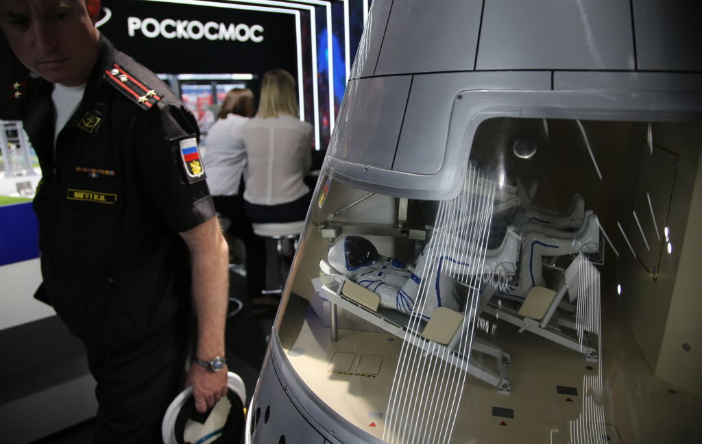 A uniformed man stands beside a display model of a spacecraft capsule at an exhibition.