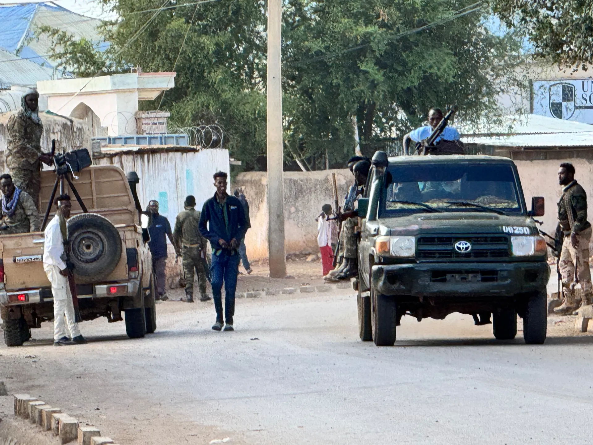 Armed men stand beside two pickup trucks on a paved street, with a pedestrian between them.