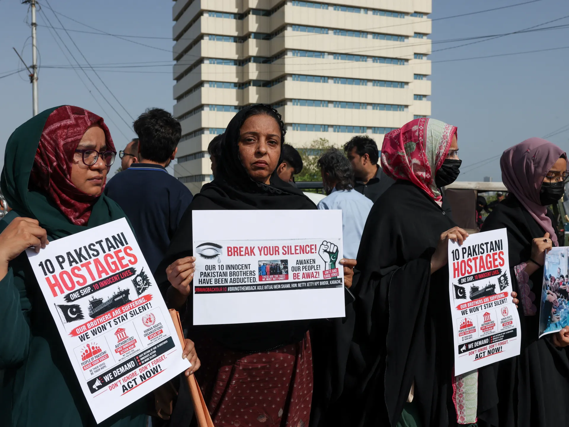 Women hold signs protesting the abduction of ten Pakistani sailors, with a high-rise building behind them.