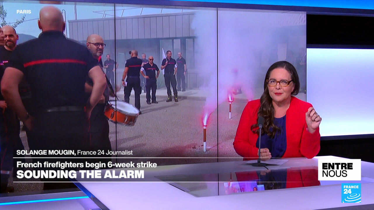 Television journalist presents footage of firefighters gathered outside amid red smoke flares