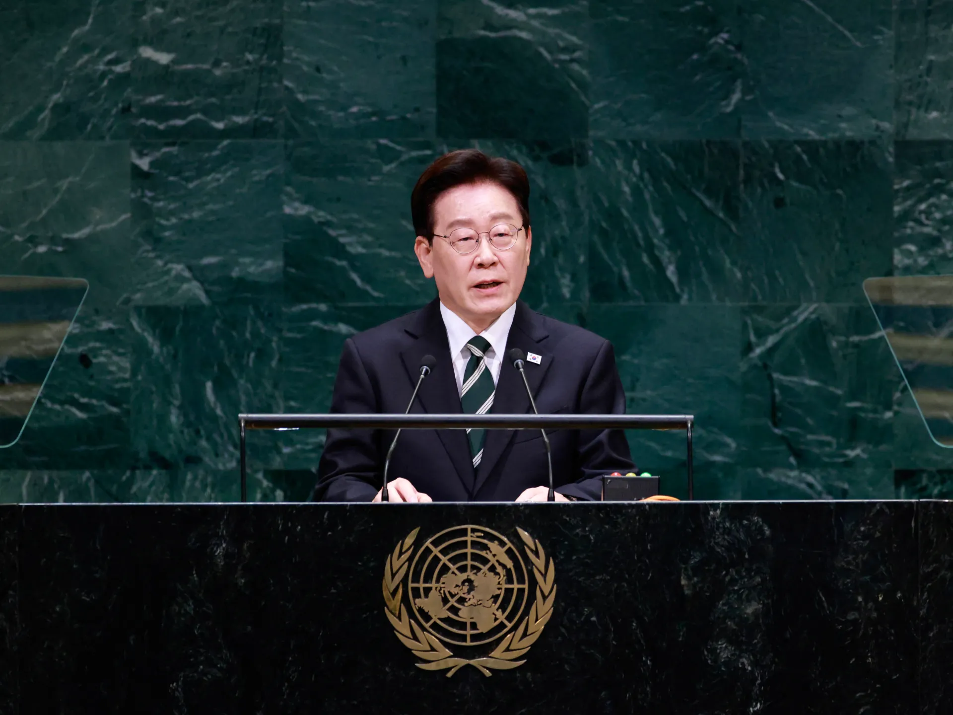 A suited man wearing glasses speaks at a lectern with microphones before a green marble wall.