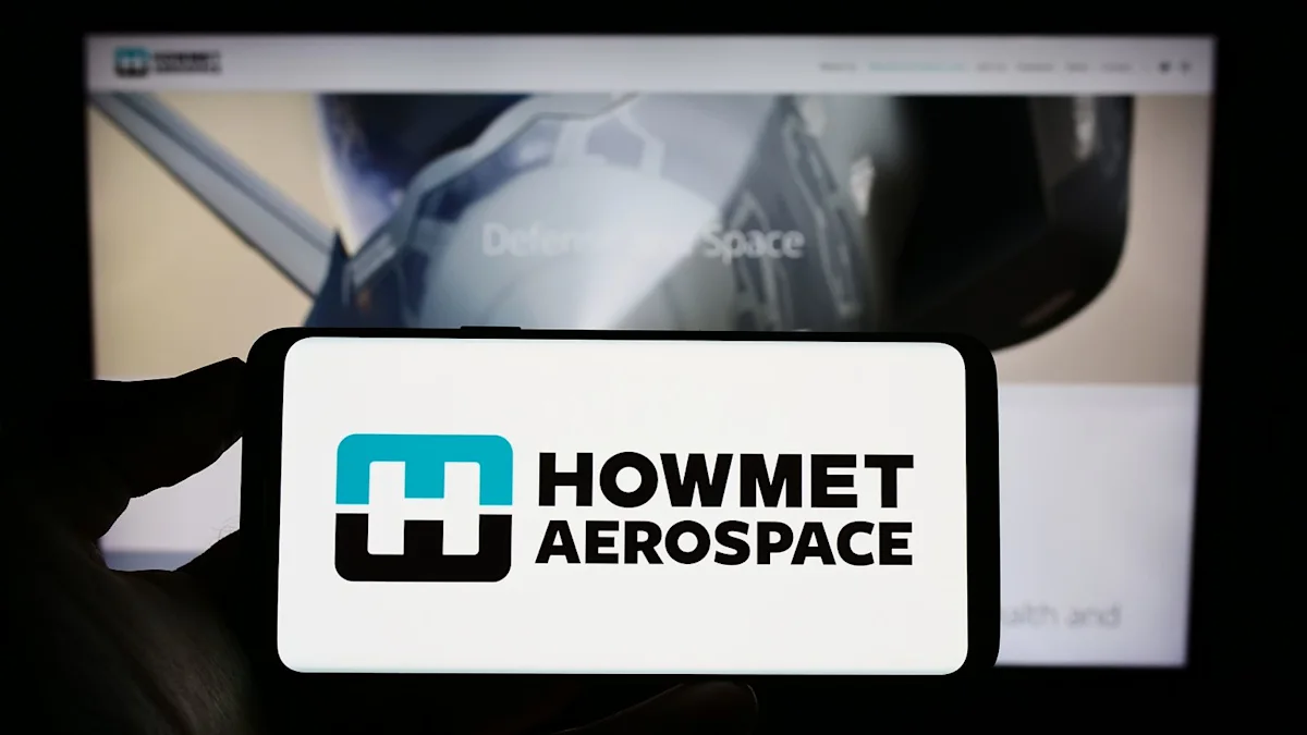 Hand holds a smartphone displaying the Howmet Aerospace logo in front of a blurred computer screen.