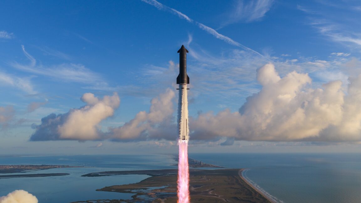 A Starship rocket ascends above coastal waters, trailing bright exhaust beneath a blue sky with clouds.