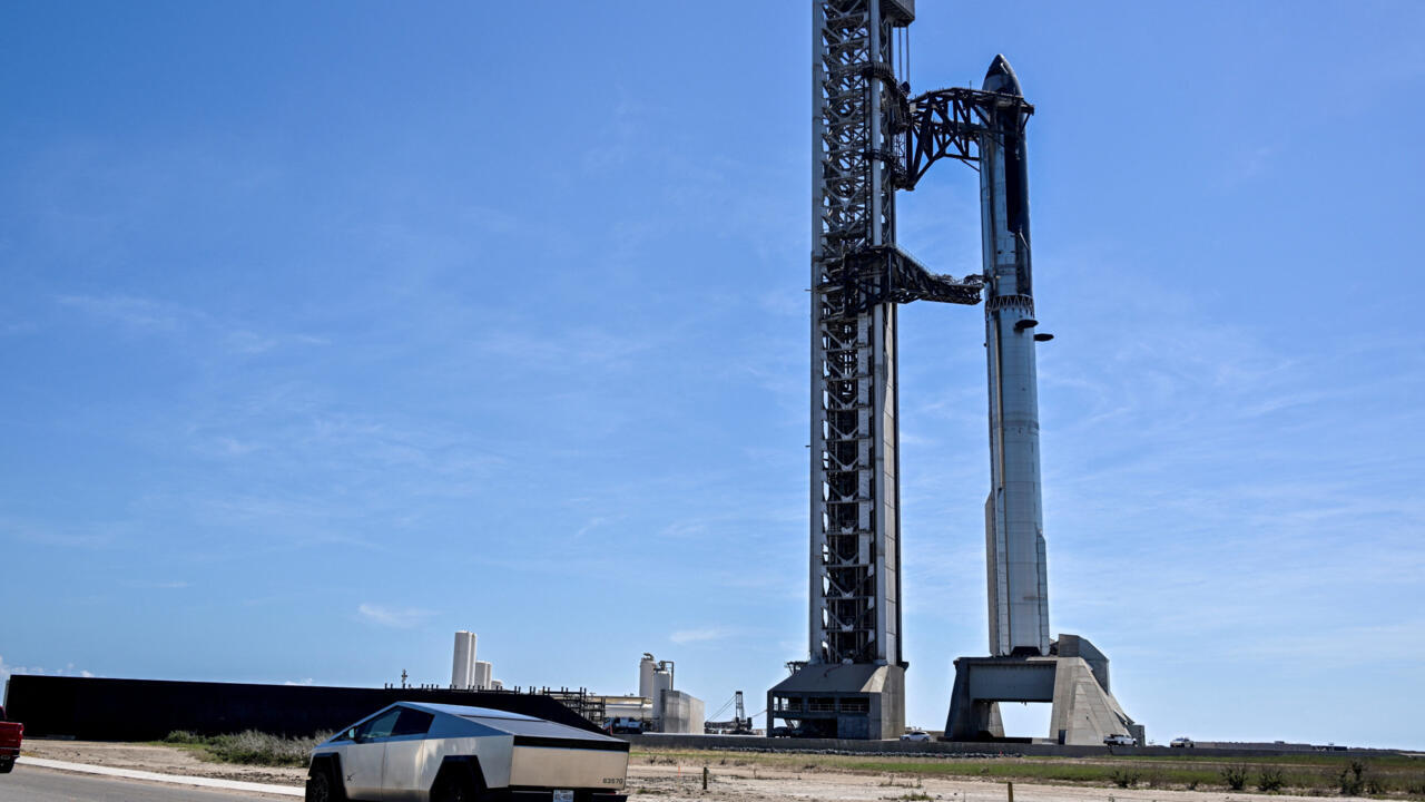 A tall rocket stands beside its launch tower under a clear blue sky, with a pickup truck in front.
