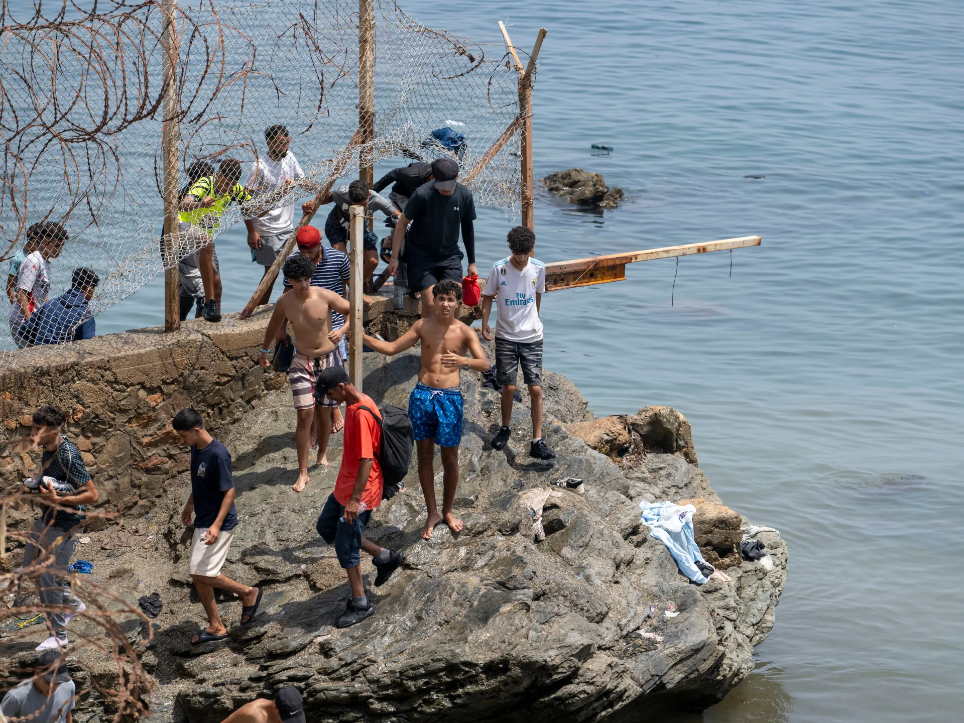 Group of young men gathered on rocky shoreline beside a damaged chain-link fence and calm water.
