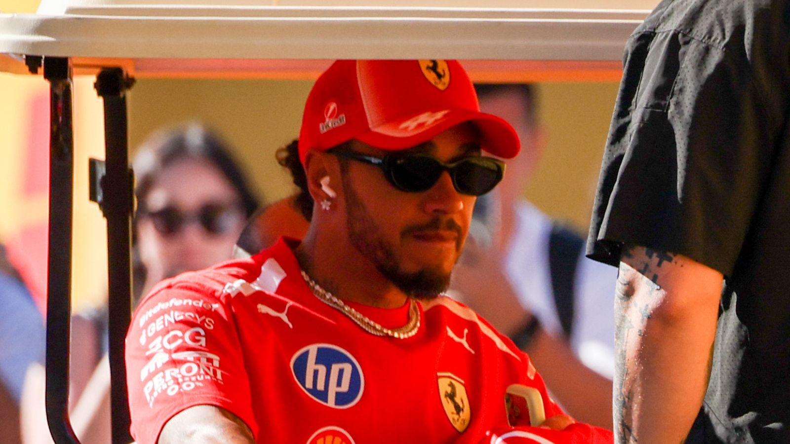Ferrari driver wearing red team clothing, sunglasses and cap sits beneath a shaded canopy