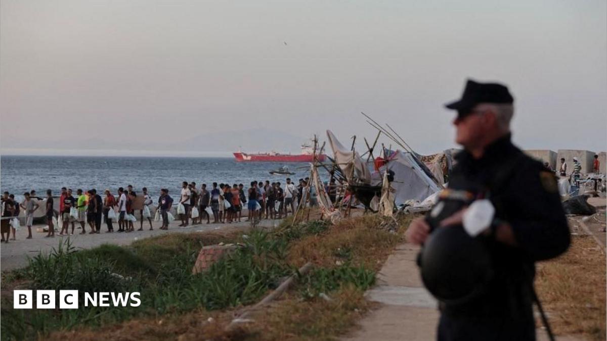 Crowd gathers along a shoreline near makeshift shelters as a uniformed officer stands in the foreground.