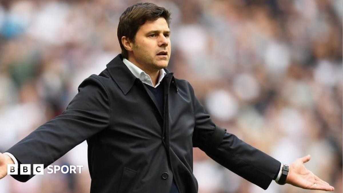 Football manager in black coat gestures with both arms extended before a blurred crowd.
