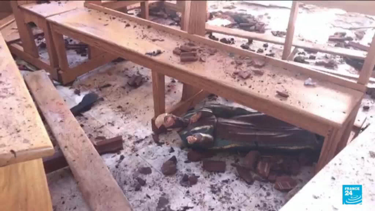 A person lies beneath a wooden table in a room with scattered debris.