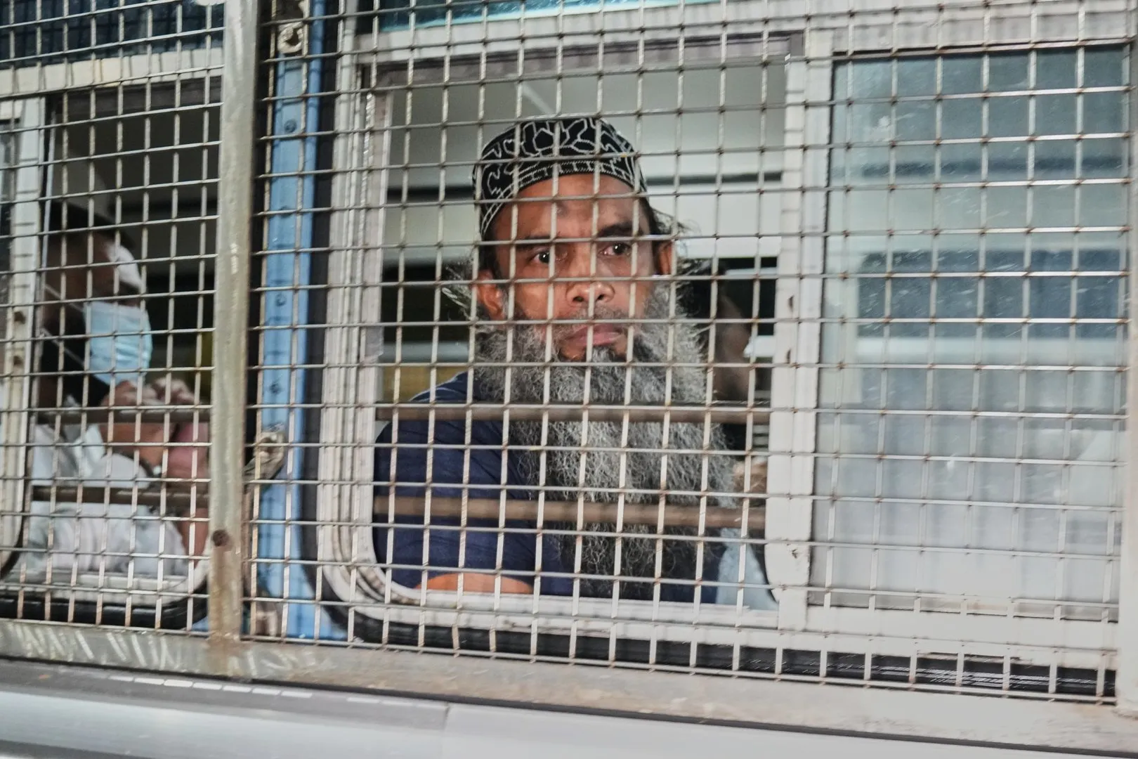 A bearded man wearing a patterned cap stands behind a metal security grille inside a building.