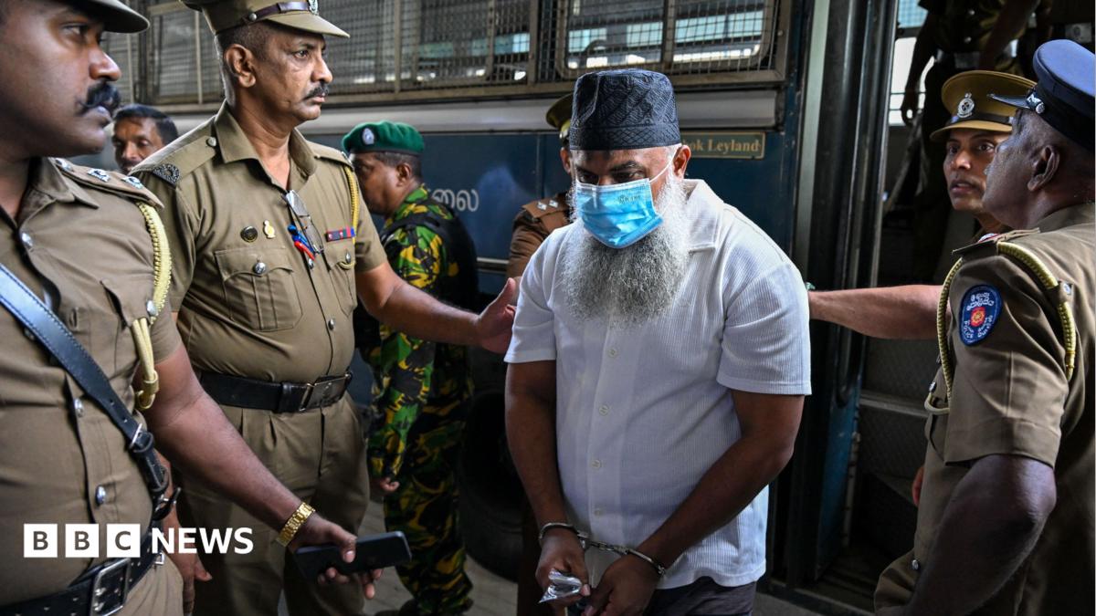 Police officers escort a masked, bearded man in handcuffs through a crowded transport vehicle.