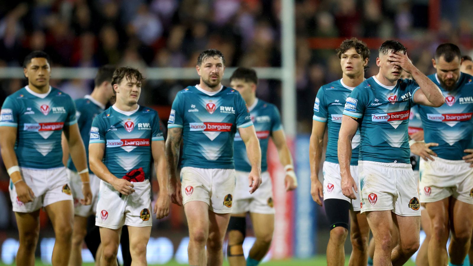 Rugby players in teal jerseys walk across the field, with one covering his face.