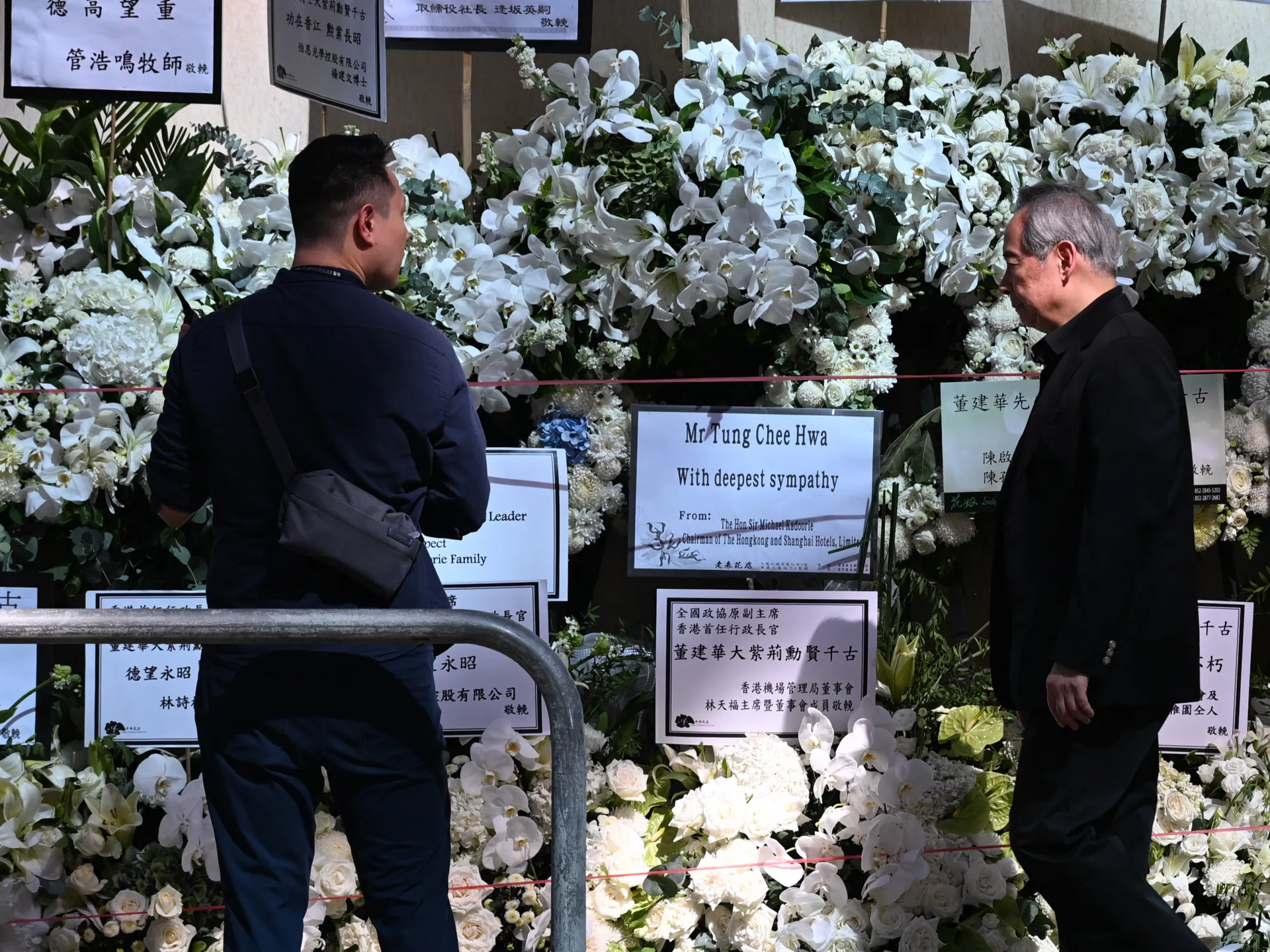 Two men view a wall of white funeral flowers and condolence wreaths behind barriers.