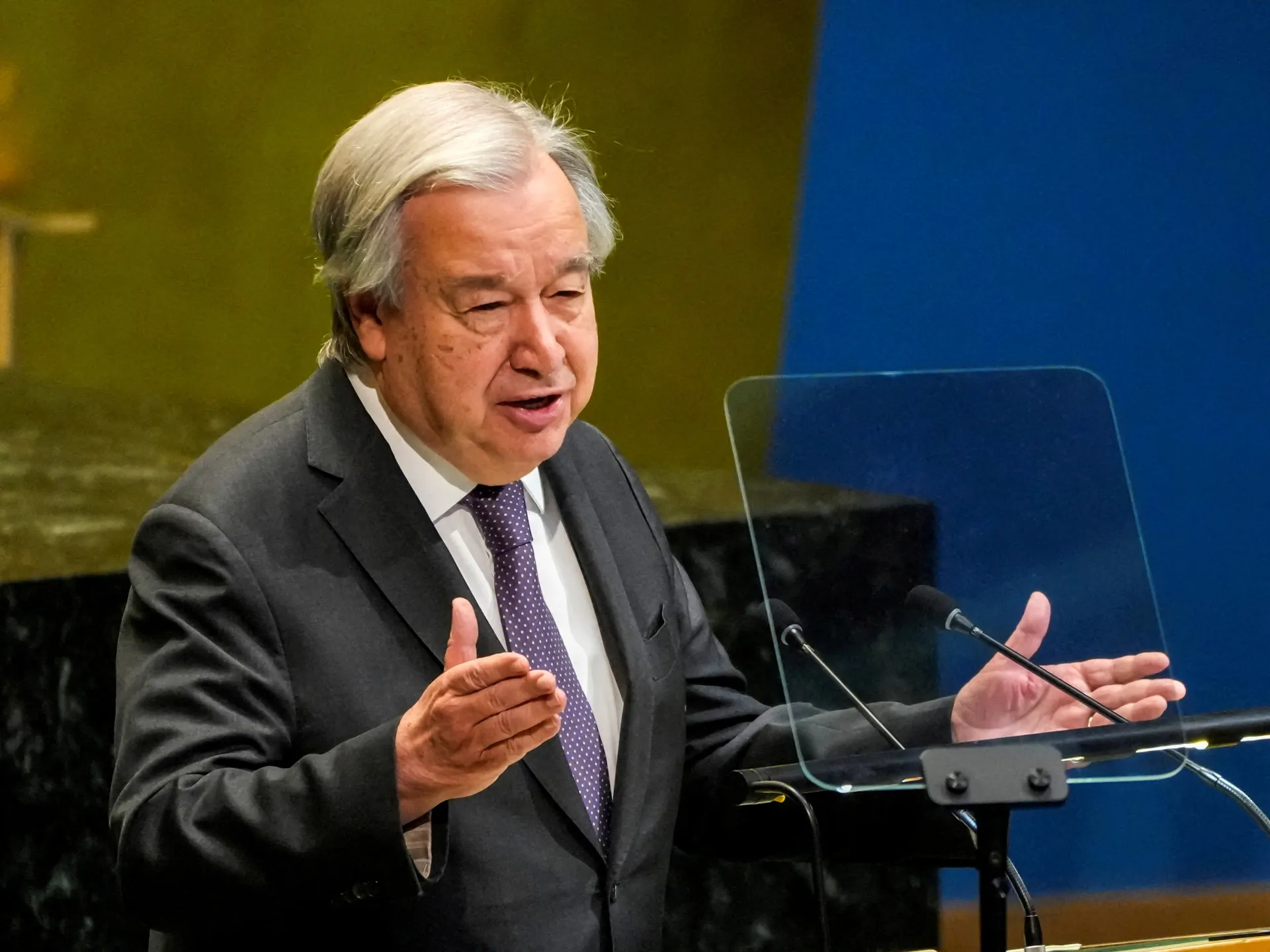 Gray-haired man in suit gestures while speaking at a lectern beside a transparent teleprompter.